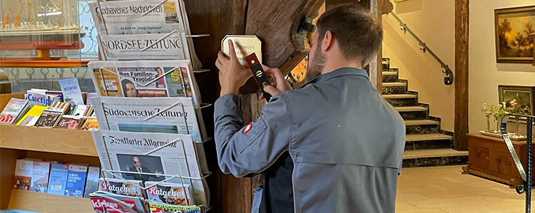Ein Mitarbeiter in grauer Arbeitskleidung führt Arbeiten an einer Holzkonstruktion in der Hotelhalle des Badhotel Sternhagen durch. Daneben steht ein Zeitungsständer mit verschiedenen Tageszeitungen.