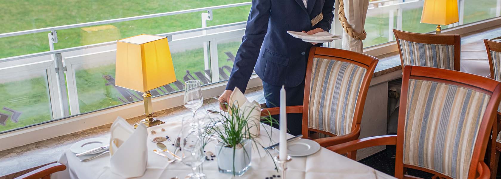 Ein Kellner in formeller Kleidung serviert Speisen an einem elegant gedeckten Tisch im Restaurant des Badhotel Sternhagen. Der Tisch ist mit feinem Geschirr, Gläsern und einem kleinen Pflanzenelement dekoriert. Im Hintergrund sind große Fenster mit Blick ins Grüne und eine stilvolle Tischlampe sichtbar.