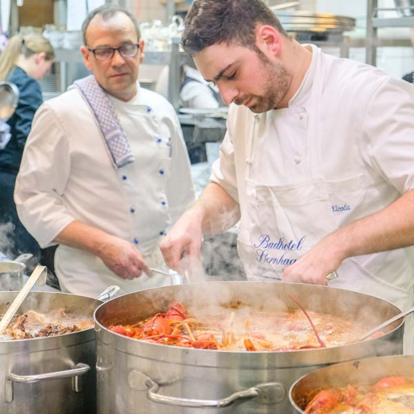 Köche im Badhotel Sternhagen bereiten in einer professionellen Küche eine Meeresfrüchte-Spezialität zu. Dampf steigt aus großen Töpfen auf, während ein junger Koch konzentriert mit einem Kochlöffel rührt. Im Hintergrund ist ein weiterer Koch sichtbar, der aufmerksam beobachtet. Die Atmosphäre vermittelt eine engagierte und kreative Küchenumgebung.