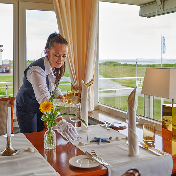 Eine Servicekraft deckt einen eleganten Tisch in einem lichtdurchfluteten Speisesaal des Badhotel Sternhagen. Auf dem Tisch befinden sich eine Vase mit einer Sonnenblume, Kerzenhalter, Servietten und dekorative Muscheln. Im Hintergrund sind große Fenster mit Blick auf die grüne Landschaft und das Meer zu sehen.