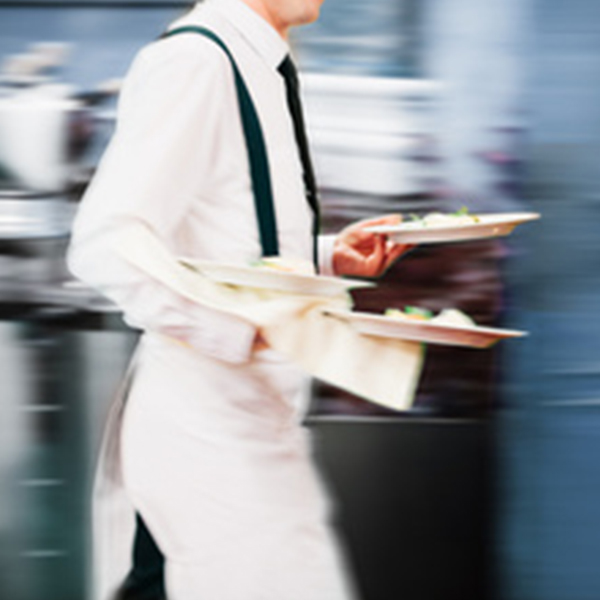 Ein Kellner in eleganter Arbeitskleidung mit Hosenträgern trägt mehrere Teller mit Speisen in einem geschäftigen Restaurant. Die Bewegung des Kellners vermittelt ein Gefühl von Dynamik und Professionalität in einem gehobenen gastronomischen Umfeld.