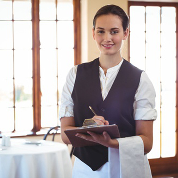 Eine Kellnerin in einem eleganten Restaurant des Badhotel Sternhagen hält ein Notizbuch und einen Stift in der Hand, während sie freundlich in die Kamera lächelt. Im Hintergrund sind große Fenster sichtbar, die viel Tageslicht hereinlassen, und ein gedeckter Tisch ist im Vordergrund zu sehen.