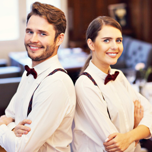 Zwei lächelnde Servicekräfte in eleganter Kleidung stehen Rücken an Rücken in einem stilvollen Hotelumfeld. Der Mann trägt eine weiße Bluse mit einer Fliege, die Frau ebenfalls in einer weißen Bluse mit einem roten Akzent. Im Hintergrund ist eine geschmackvoll eingerichtete Lounge zu sehen.
