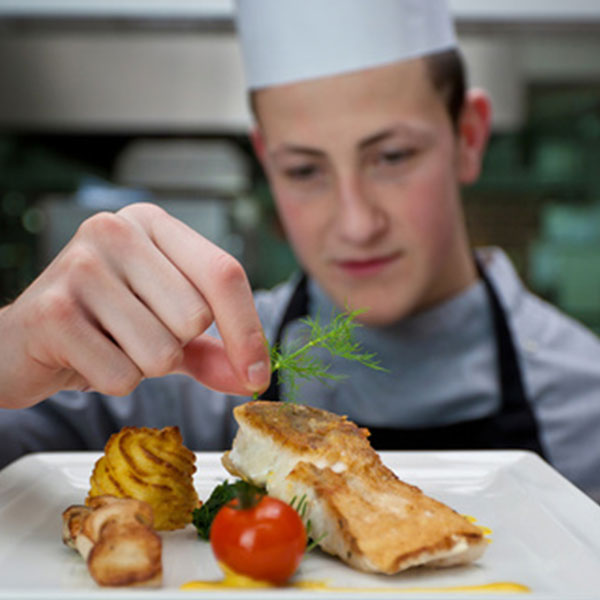 Ein junger Koch dekoriert ein kunstvoll angerichtetes Gericht mit gebratenem Fisch, Beilagen und einer frischen Kräuter garnitur. Im Hintergrund sind Küchenelemente zu erkennen, die die professionelle Atmosphäre des Badhotel Sternhagen unterstreichen.