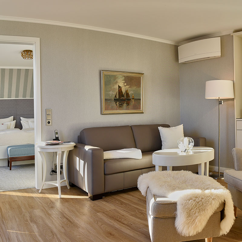 Elegantes Zimmer im Badhotel Sternhagen mit komfortablem Sofa, stilvollem Tisch und kunstvollem Gemälde an der Wand. Die warmen Holzböden und die geschmackvolle Einrichtung schaffen eine einladende Atmosphäre.