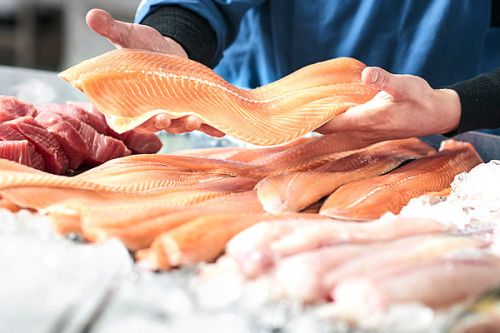 Frischer Lachs und andere Fischsorten liegen auf Eis, während eine Hand ein Stück Lachs präsentiert. Der Hintergrund zeigt einen professionellen Fischverkäufer in einem blauen Outfit.